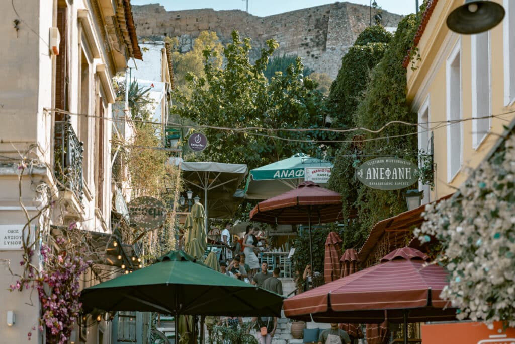 Bustling café-lined steps on Mnisikleous Street in Plaka (Plaka stairs) with colorful umbrellas and signs, framed by greenery and the Acropolis wall towering above, showing the lively atmosphere you’ll encounter during a day in Athens.