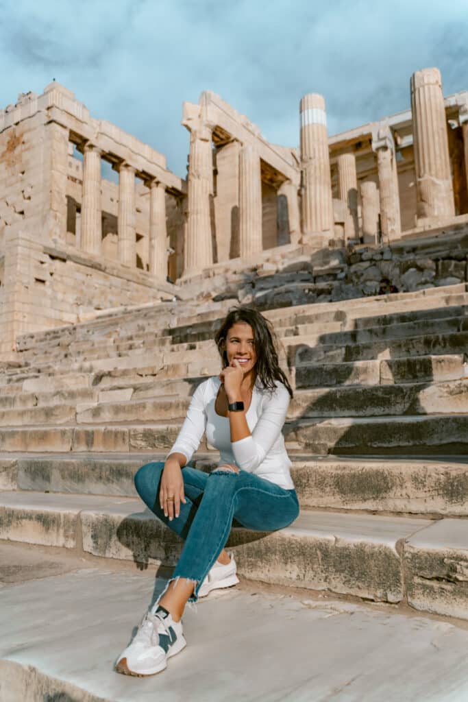 Me sitting on the ancient stone steps of the Acropolis in Athens, smiling with the towering columns of the Propylaea behind her on a sunny day.