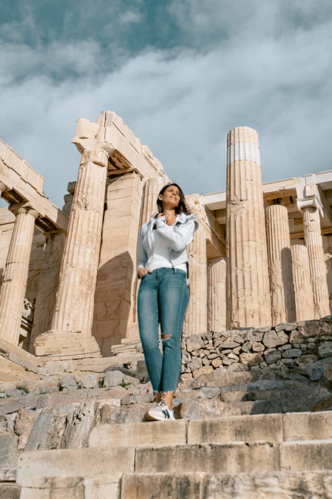 Me tanding confidently on the stone steps of the Acropolis in Athens, looking up with her hand near her neck, surrounded by tall marble columns under a bright sky.