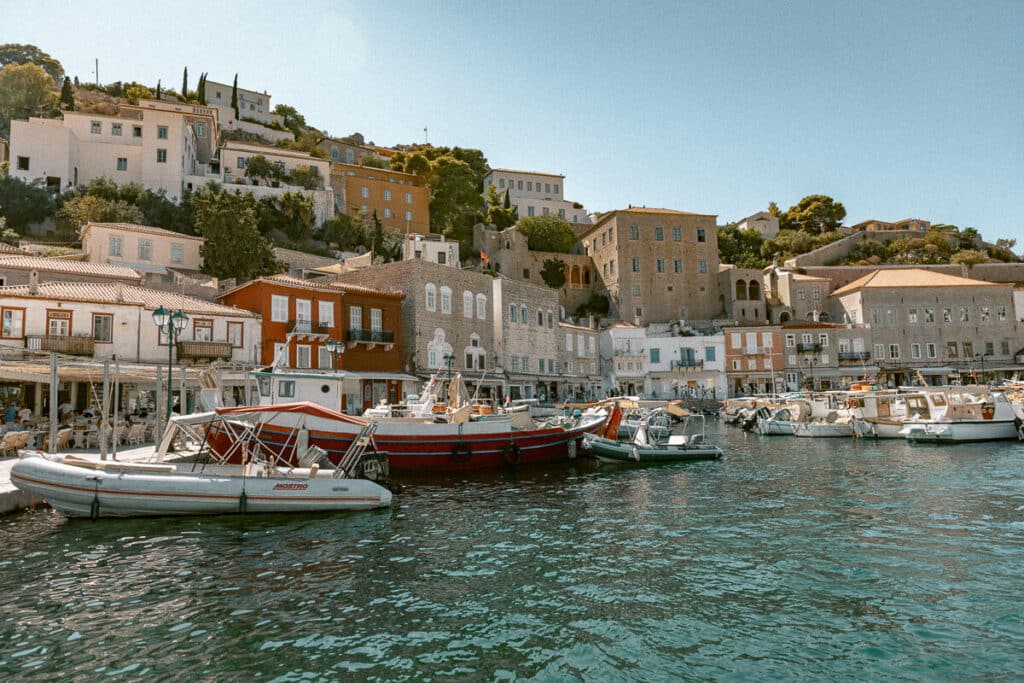 Colorful boats docked along the waterfront in Hydra, Greece, with traditional stone houses climbing the hillside behind the harbor.