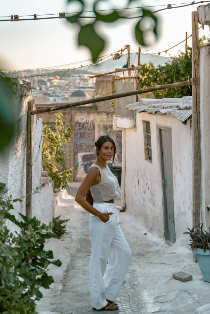 Me in white linen pants standing in a narrow Anafiotika alley with whitewashed walls and views of Athens in the background, capturing the island vibe in the city.