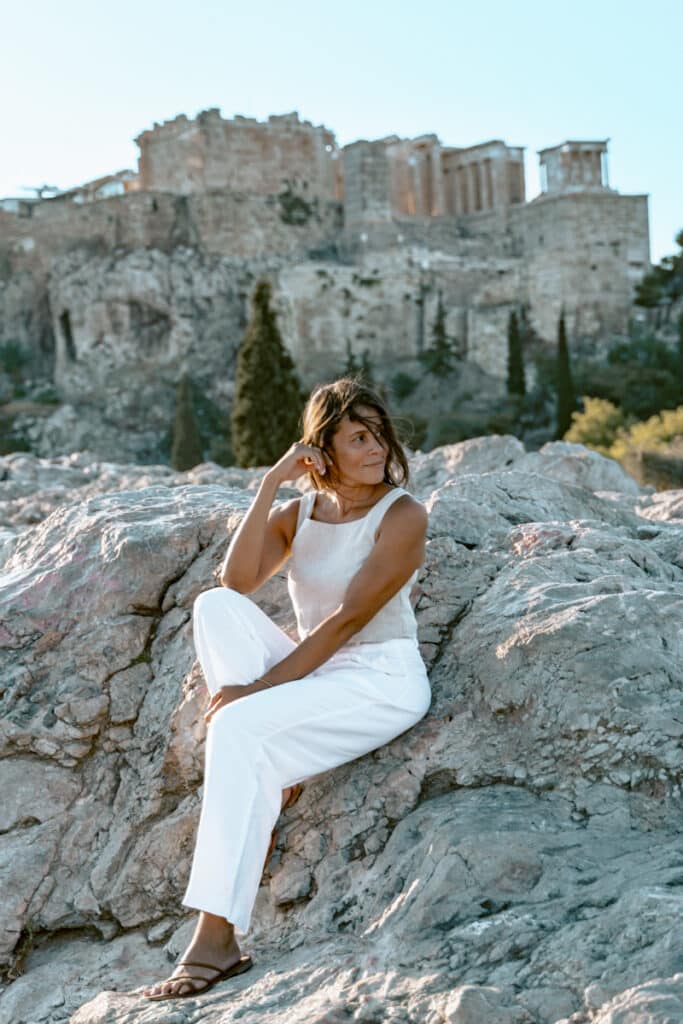 Me in white linen sitting on rocky outcrops with the Acropolis ruins rising in the background from Filopappou hill, a quiet spot for reflection during a visit to Athens.