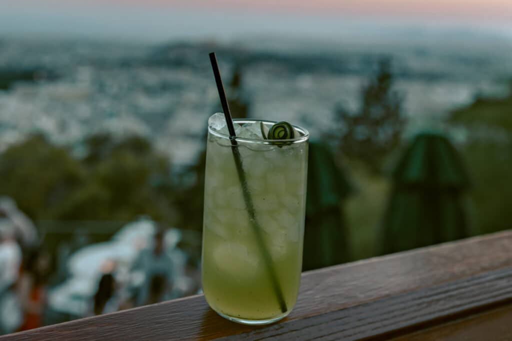 A chilled cocktail with cucumber garnish on a bar ledge at Prasini Tenta, with Athens city views blurred in the background, representing rooftop dining in Athens itineraries.