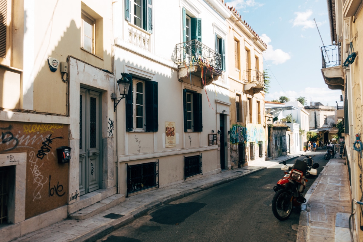 A narrow street in Athens with pastel-colored buildings, graffiti on the walls, balconies with shutters, and a parked motorbike. The quiet scene captures the charm of exploring local neighborhoods on an Athens 2 day itinerary, where everyday life and history blend together.