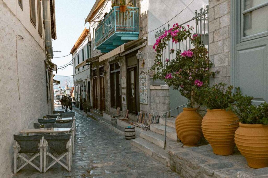 Narrow cobblestone alley in Hydra with outdoor café tables, bougainvillea, and a donkey in the distance.