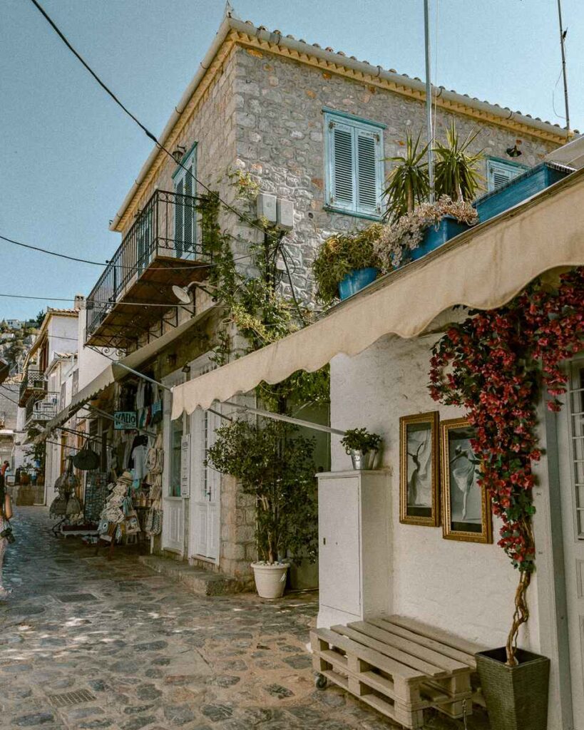 Charming stone house with blue shutters and flowers along a cobblestone street in Hydra, a picturesque stop.