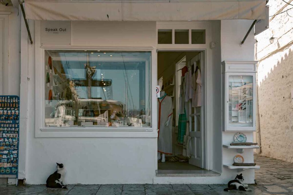 Whitewashed boutique storefront with cats lounging outside in Hydra’s harbor, a quiet street scene.