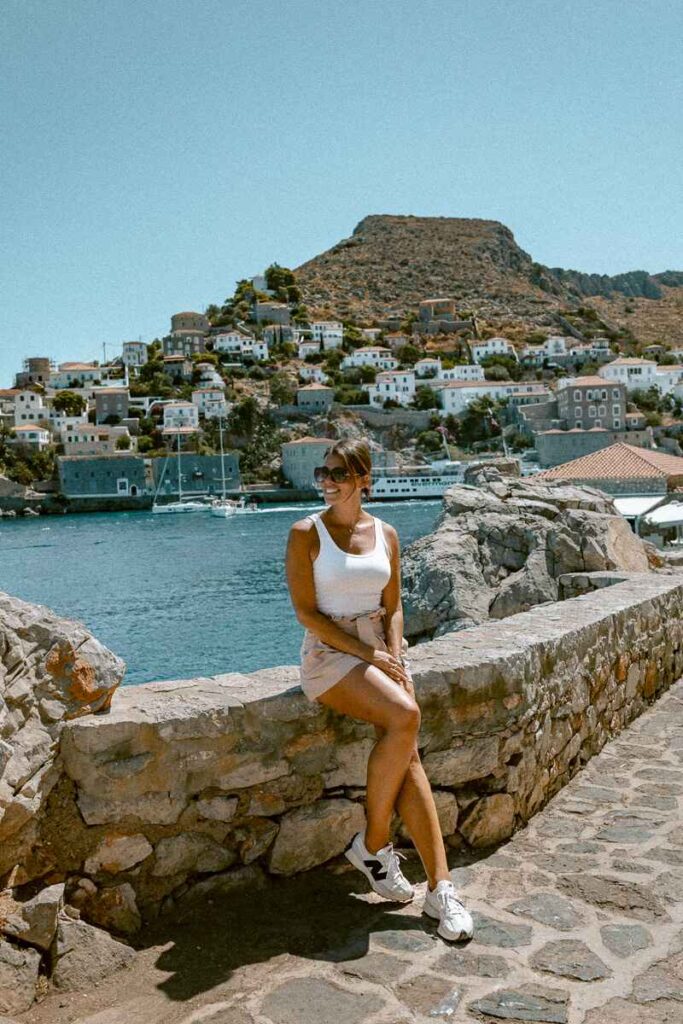 Me sitting on a stone wall with Hydra’s harbor and hillside houses in the background, capturing memories from an Athens to Hydra day trip.
