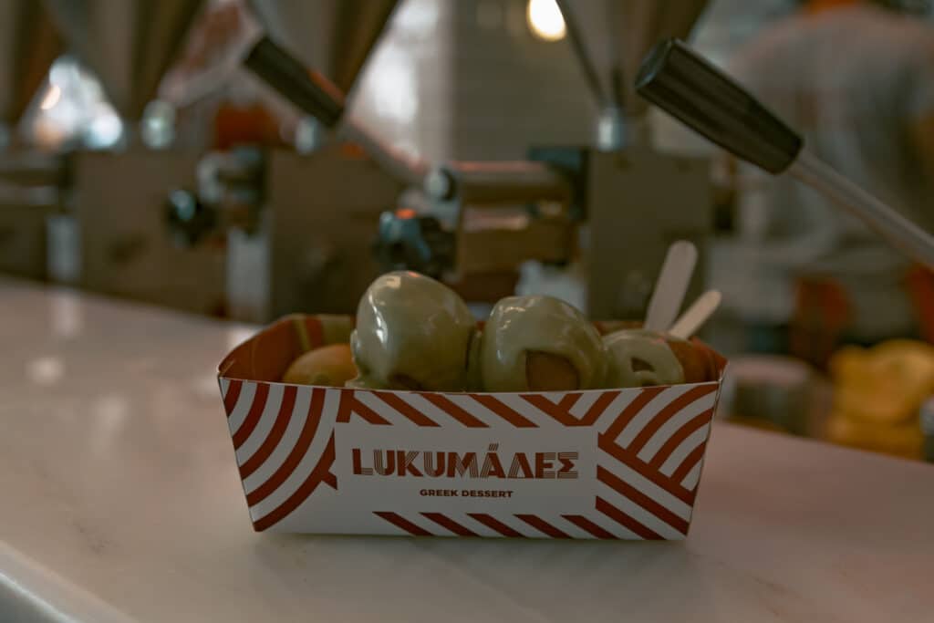 A close-up of a serving of loukoumades, traditional Greek doughnuts, covered in a creamy green pistachio sauce. The dessert is placed in a red and white striped container labeled “Lukumádes Greek Dessert,” sitting on a counter inside a shop in Athens.