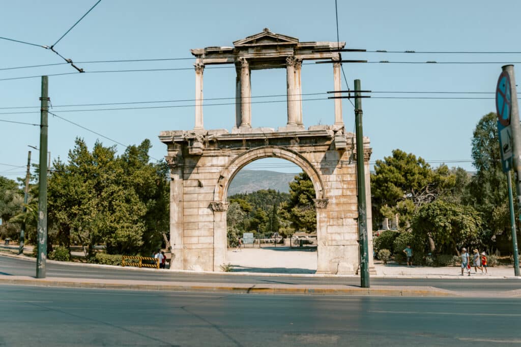 Ancient stone arch standing along a busy road in Athens with trees and mountains in the background. A landmark often passed by visitors exploring the city.