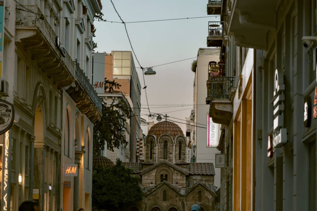 Crowded shopping street in central Athens with a small Byzantine church framed by modern buildings. Shows the lively, safe atmosphere in tourist areas to showcase the question is Athens safe.