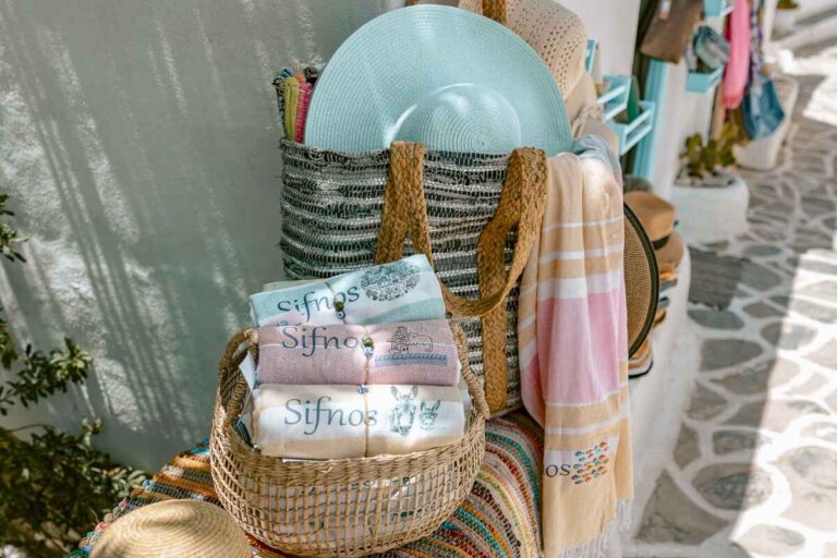 Basket of embroidered towels with “Sifnos” designs, alongside hats and bags outside a souvenir shop, a popular pick for what to buy in Greece.