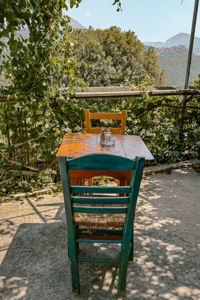 Rustic wooden table and chairs at a leafy taverna terrace in the mountains of Ikaria Island, Greece, offering a peaceful spot for enjoying local food surrounded by nature.