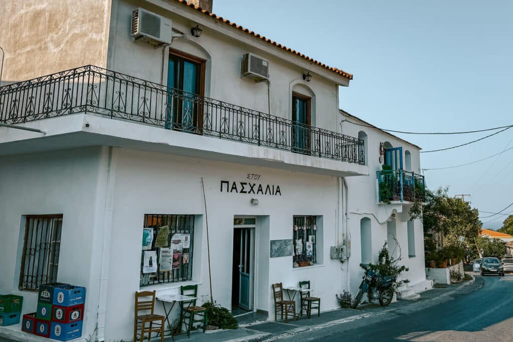 Traditional white building with blue doors and iron balconies in one of Ikaria’s charming villages, with chairs set outside for locals to relax.