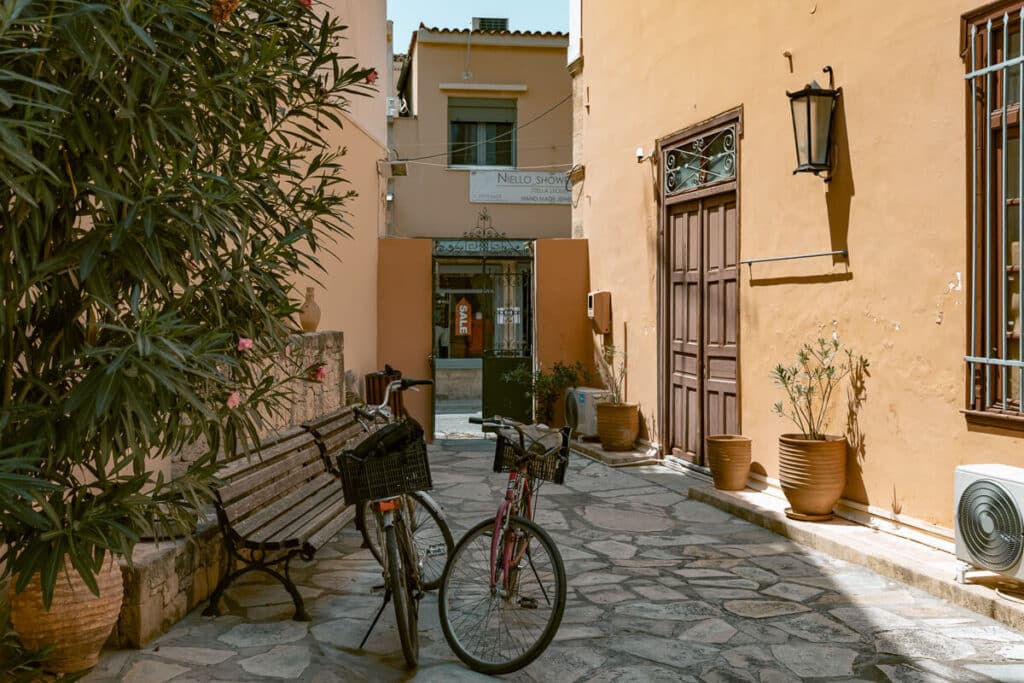 Quiet alley in Aegina with pastel-colored walls, bicycles parked beside a bench, and potted plants adding charm to the walkway — a hidden corner often missed on island day trips from Athens.