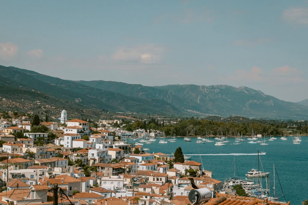 Elevated view of Poros with red-roofed houses overlooking the bay filled with sailboats and a backdrop of mountains.