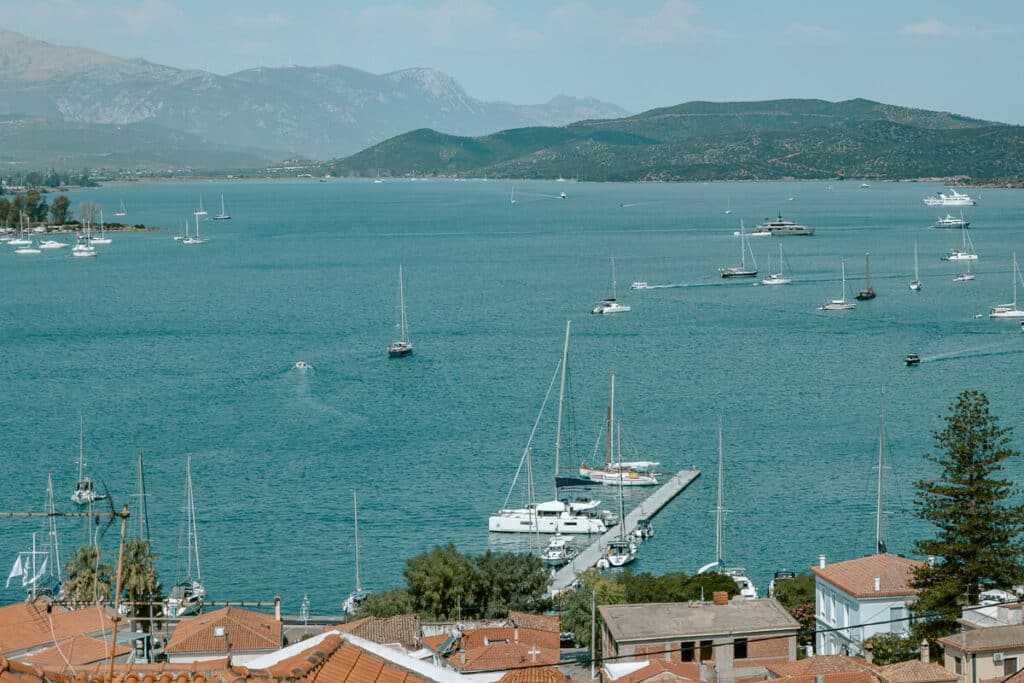 View of Poros harbor with sailboats and yachts anchored in the turquoise water, framed by terracotta rooftops and mountains in the distance.