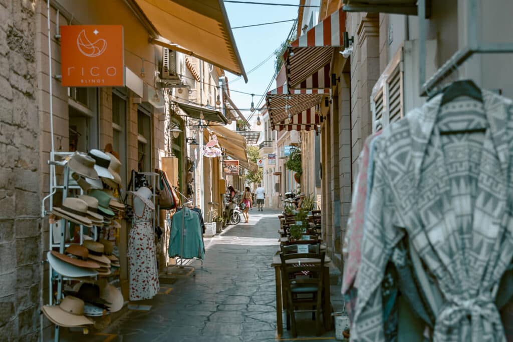 Narrow stone alleyway in Aegina Town with colorful awnings, hats, and dresses on display at shops.