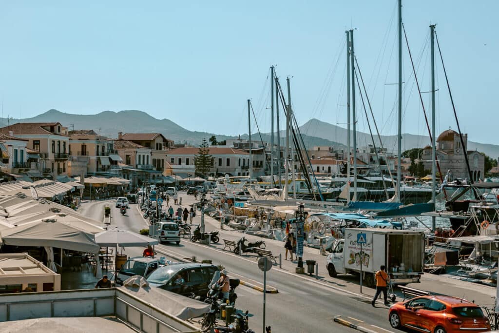 Busy harbor scene in Aegina with sailboats, waterfront cafés, and mountains in the background, one of the top island day trips from Athens.