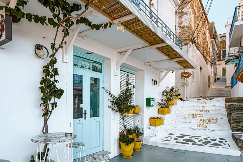 White steps in Batsi, Andros, with yellow planters, blue doors, cafe signs, and painted text reading The G Corner Batsi Andros 2026.
