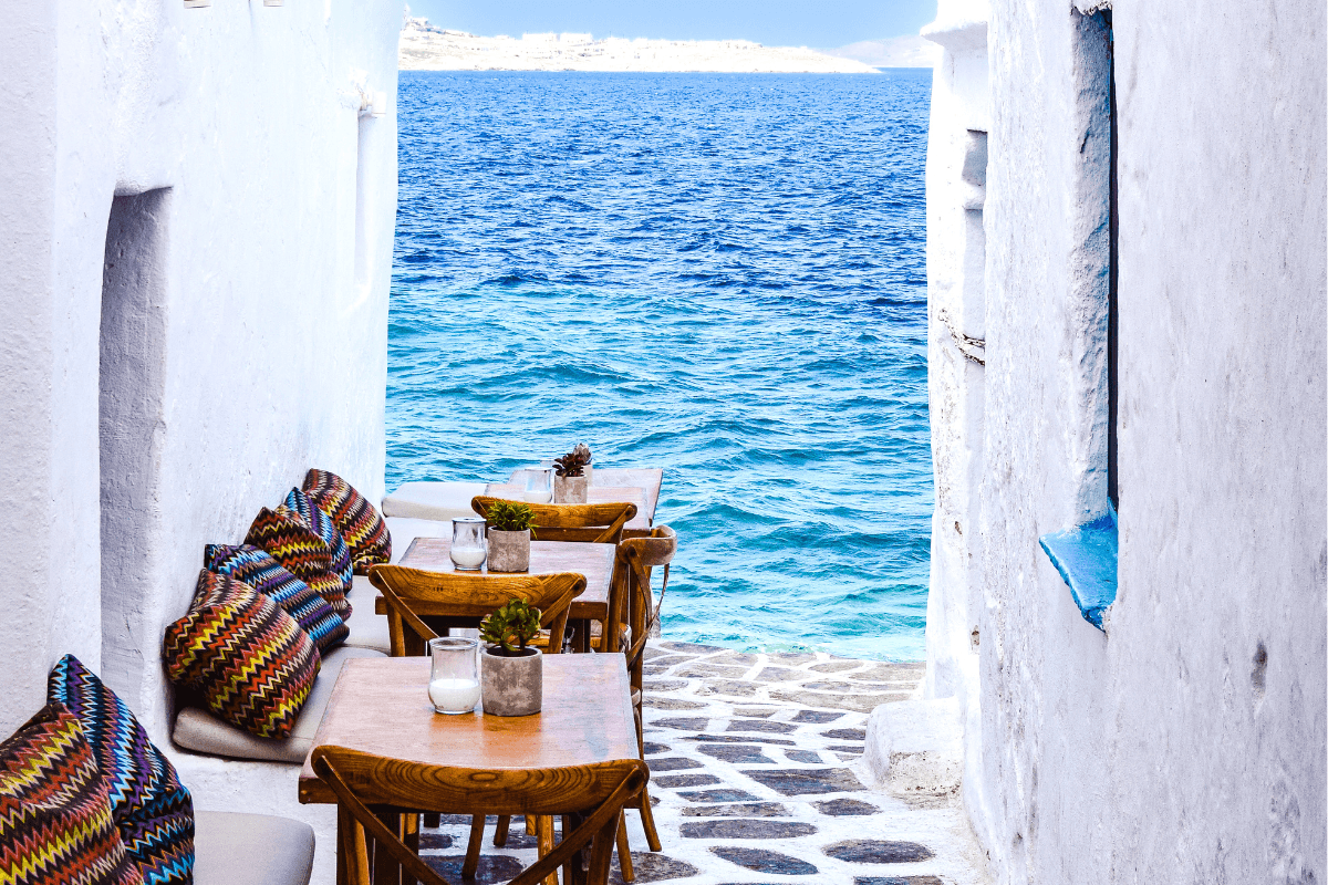 Seaside café with wooden tables and colorful cushions between whitewashed walls overlooking the bright blue Aegean Sea, featured in a collection of Greek island captions.