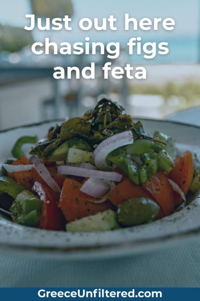 A fresh Greek salad topped with feta, tomatoes, cucumbers, onions, and herbs sits on a table with the sea in the background and the phrase “Just out here chasing figs and feta.”
