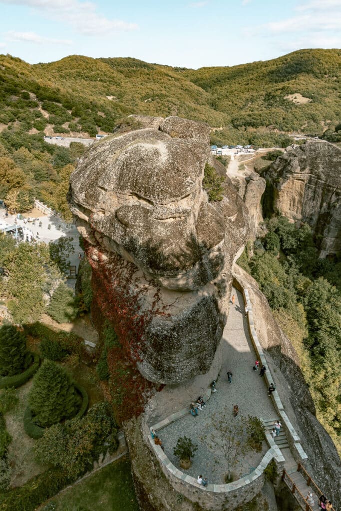 Looking down from the cliffs of Meteora at a winding stone pathway, with visitors exploring the epic rock formations and gardens below perfect views for any Meteora guide.