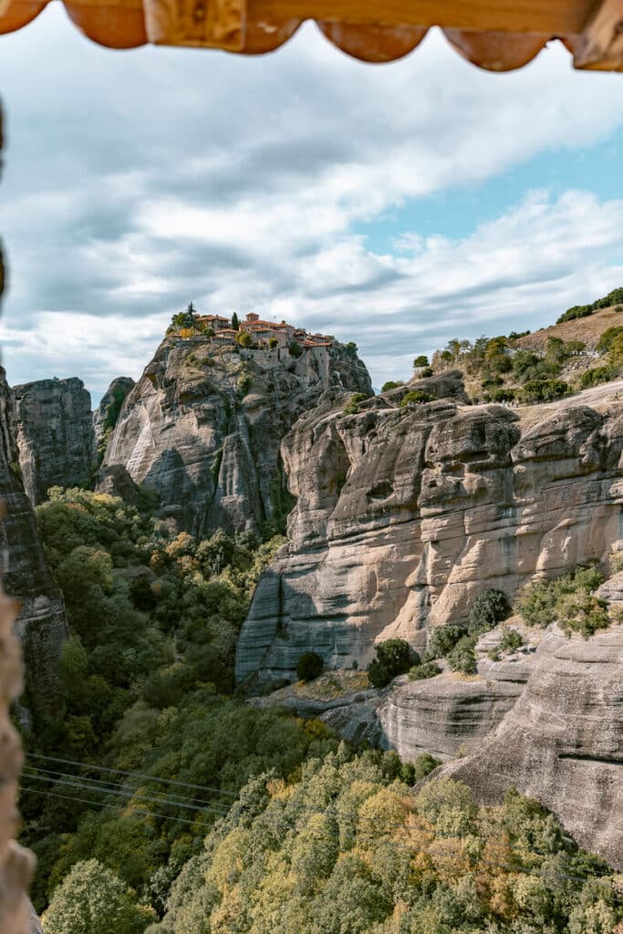 Dramatic view of another Meteora monastery balanced on a rocky peak, seen through a rooftop overhang, with striking clouds and landscape in the background.