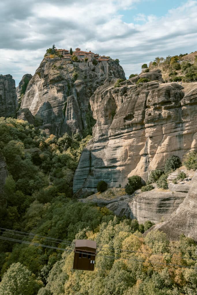 Dramatic view of another Meteora monastery balanced on a rocky peak, seen through a rooftop overhang, with striking clouds and landscape in the background.