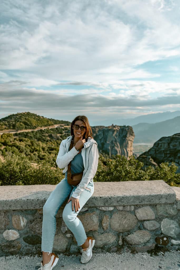 Me in casual clothes sitting on a stone wall, smiling with Meteora’s famous cliffs and valleys in the background a must see on any Meteora guide.