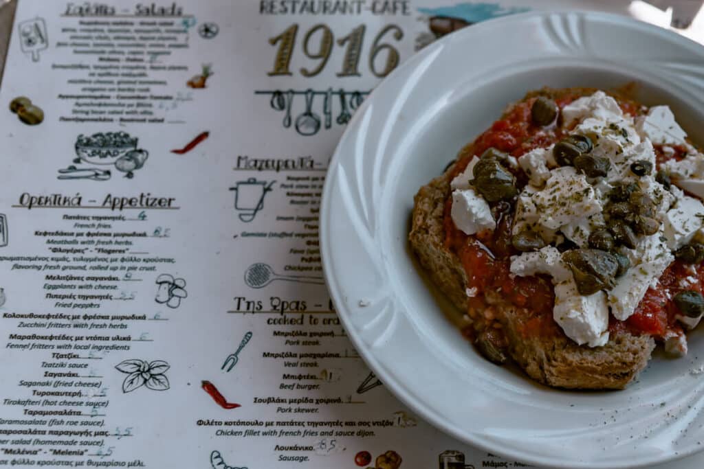 A Greek dakos salad served on a rustic menu background at a Serifos café, topped with feta cheese, tomatoes, and capers.