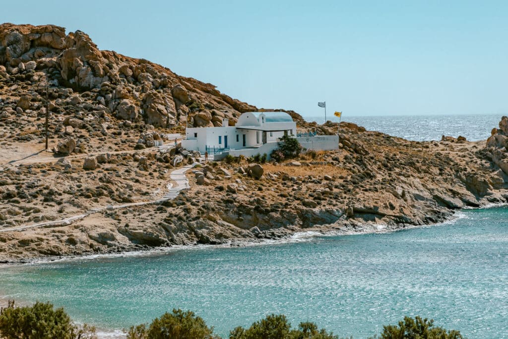 Charming white chapel on a rocky coastline in Serifos with Greek flags fluttering above the blue-green sea.