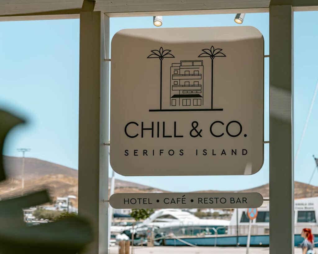 Sign for Chill & Co Serifos Island, a stylish hotel, café, and bar located by the marina with Cycladic hills in the background.
