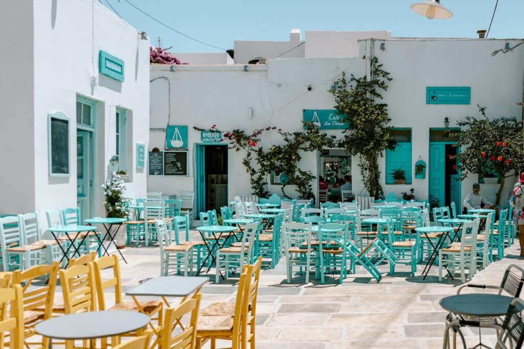 A charming Greek café in Serifos Chora with turquoise chairs and tables set in a sunlit courtyard, surrounded by whitewashed buildings and climbing vines. One of the top things to do in Serifos.