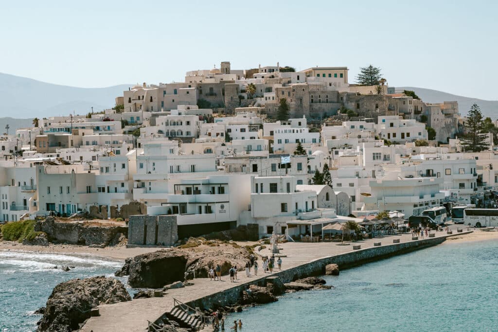 Whitewashed buildings climbing the hillside of Chora, Naxos, above a coastal walkway and turquoise Aegean water.