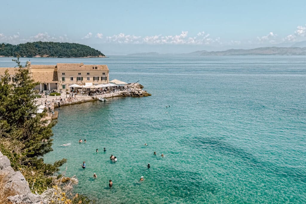 Swimmers in turquoise water near a seaside restaurant in Corfu, Greece.