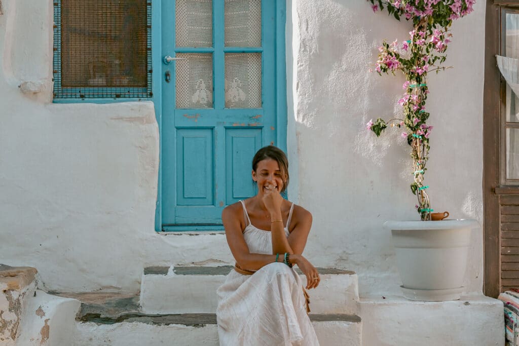 Woman sitting in front of a blue door smiling in the sun, surrounded by Cycladic architecture, that is a perfect backdrop for solo female travel Greece.