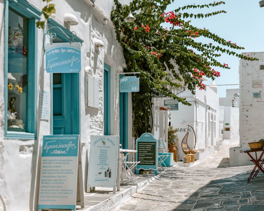 A sunlit alley in Sifnos, Greece lined with traditional whitewashed shops featuring turquoise signs, blooming bougainvillea, and outdoor seating — a picturesque spot that captures the island’s charm and slow-travel feel.