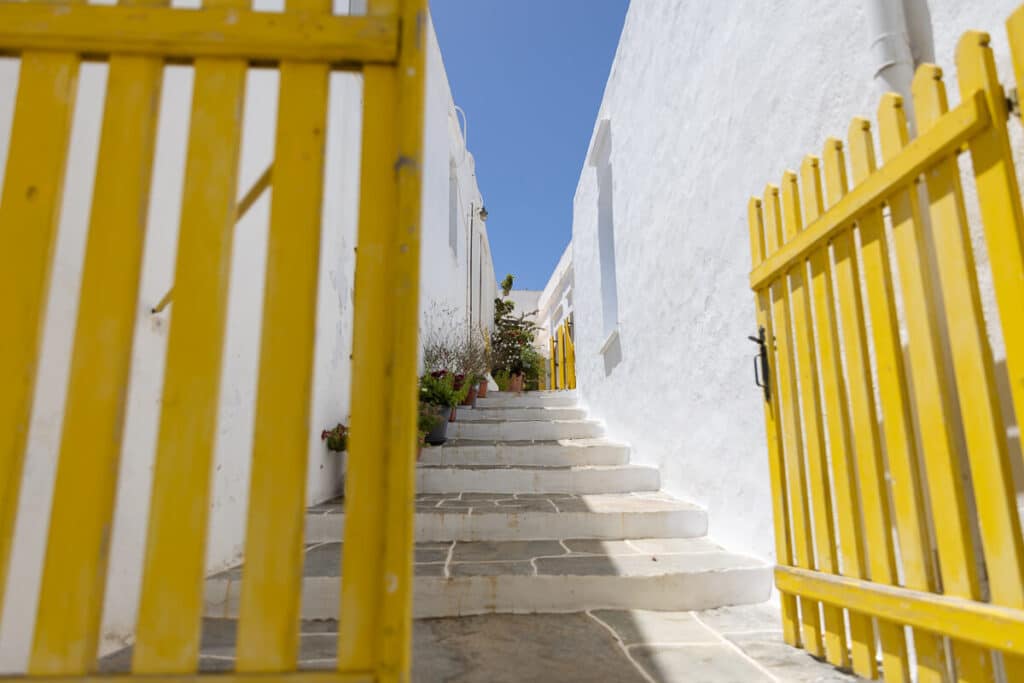A white stone staircase leading up between traditional homes in Sifnos, framed by a bold yellow gate and potted plants, a vibrant pop of color in a peaceful Cycladic alleyway.