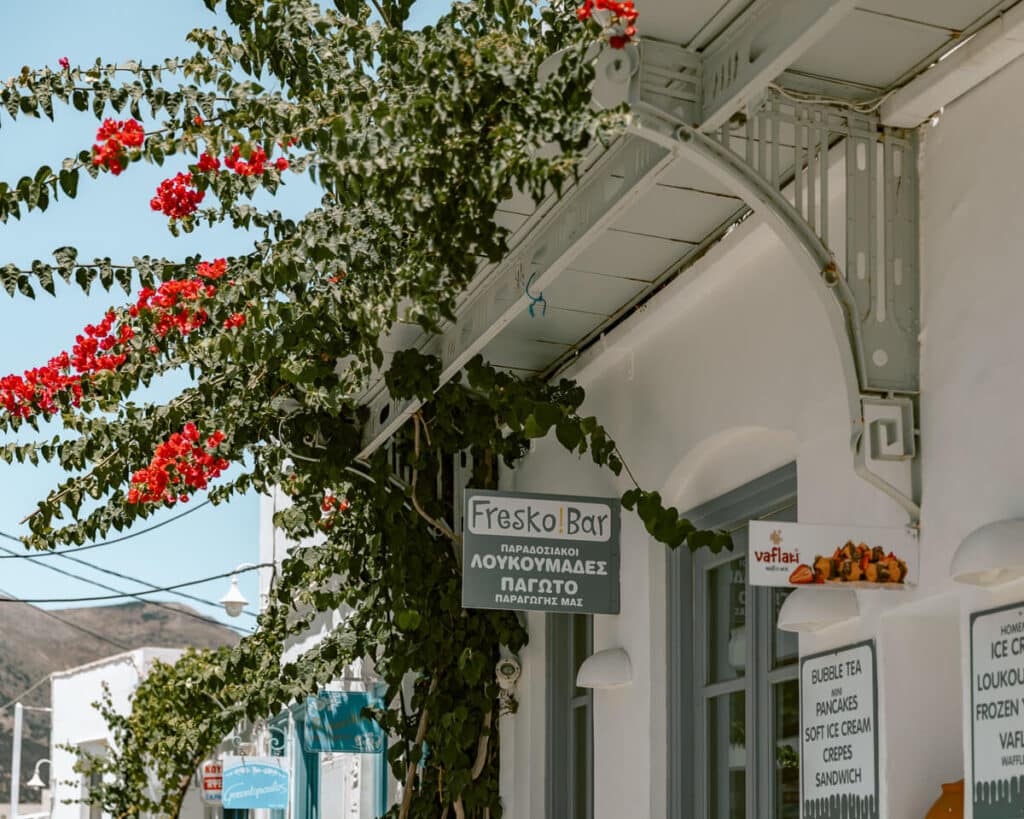 The exterior of Fresko Bar in Sifnos, Greece with a cluster of bright red bougainvillea overhead and hand-painted signs offering loukoumades and soft ice cream — a cozy local treat stop on the island.