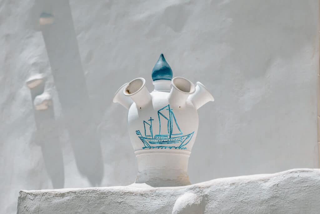 A white pottery jug with four spouts and a blue painted sailboat, placed against a whitewashed wall in Sifnos, Greece.