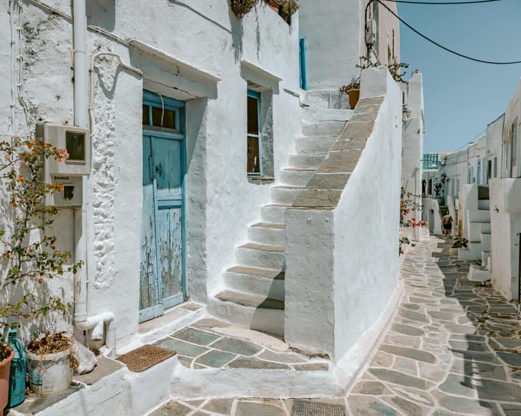 A peaceful whitewashed alley in Sifnos featuring a worn blue wooden door, stone steps, and traditional Cycladic homes.
