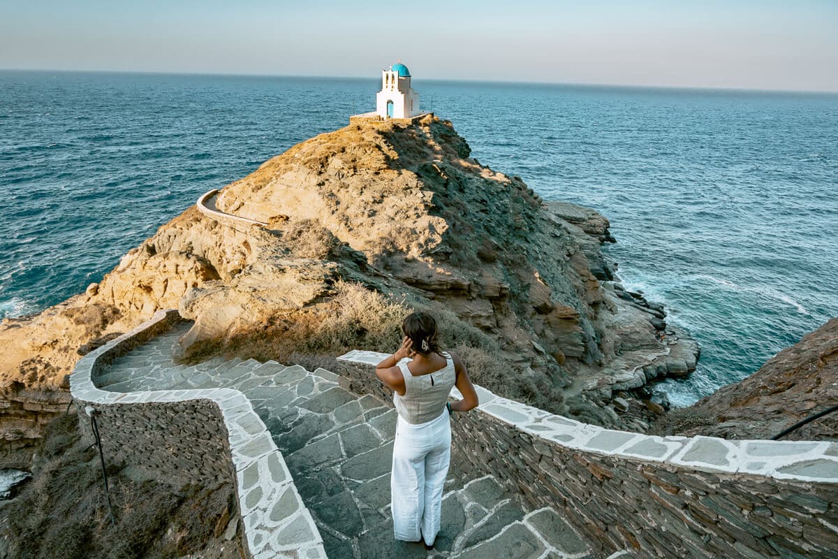 Me standing on a winding stone path overlooking the cliffside Church of the Seven Martyrs, surrounded by the deep blue Aegean Sea — a moment that visually answers the question, is Sifnos worth visiting.
