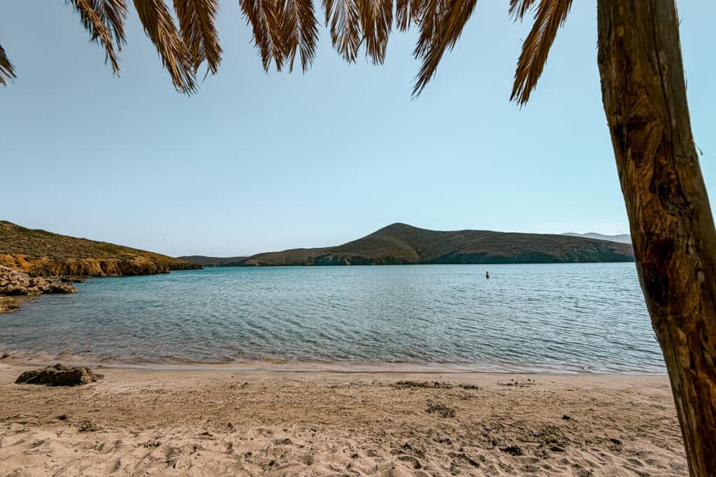 Calm turquoise water at Steno Beach in Astypalaia, with a sandy shoreline, palm leaves overhead, and dry hills across the bay.