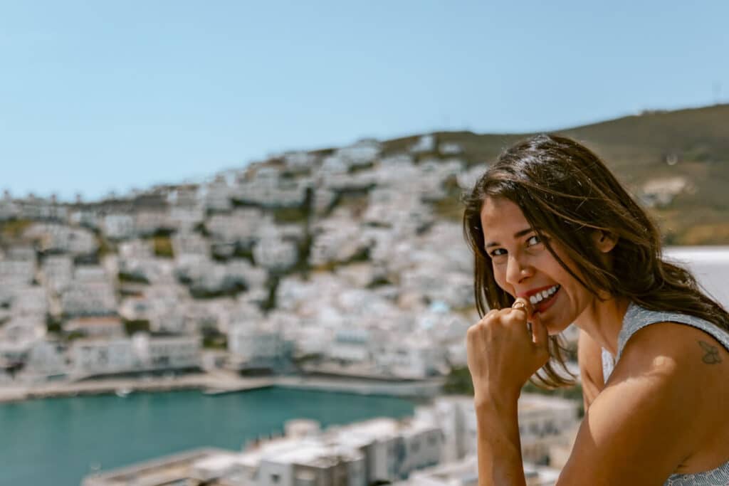 Me smiling woman leaning on a balcony railing with the iconic white homes of Astypalaia blurred in the background