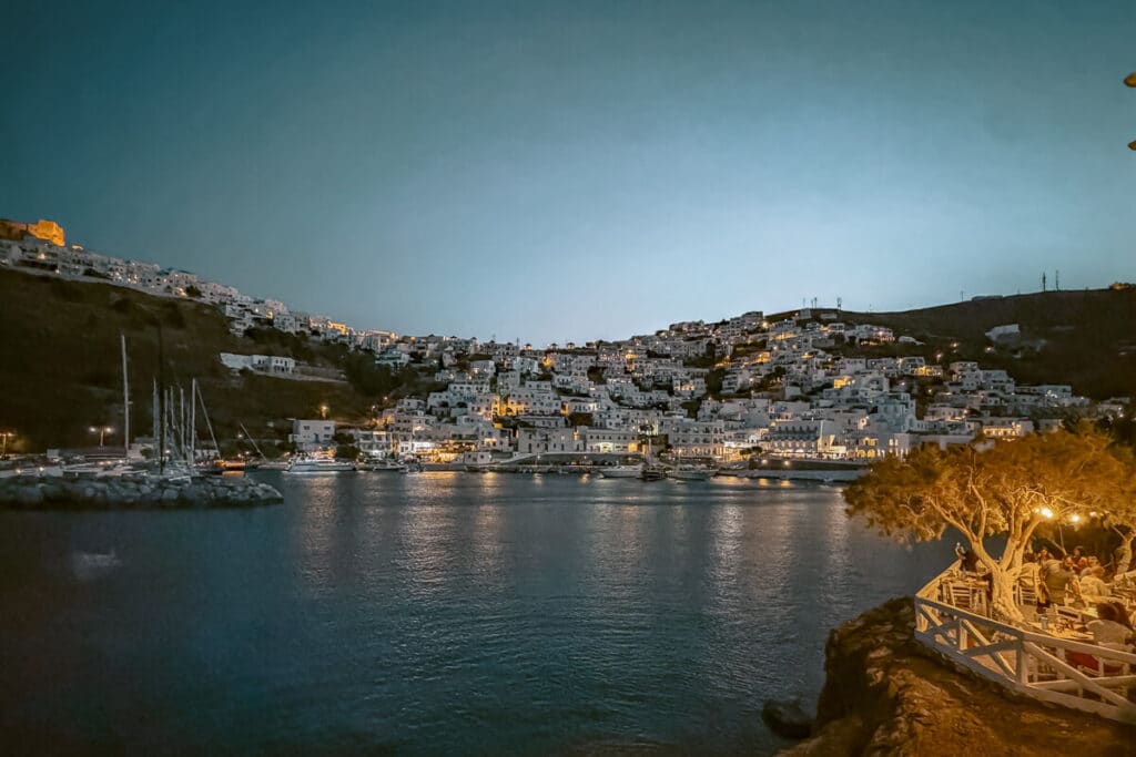 Evening view from AntiKastro in Astypalaia, with Chora’s whitewashed houses glowing across the harbor and diners seated on a terrace by the water.