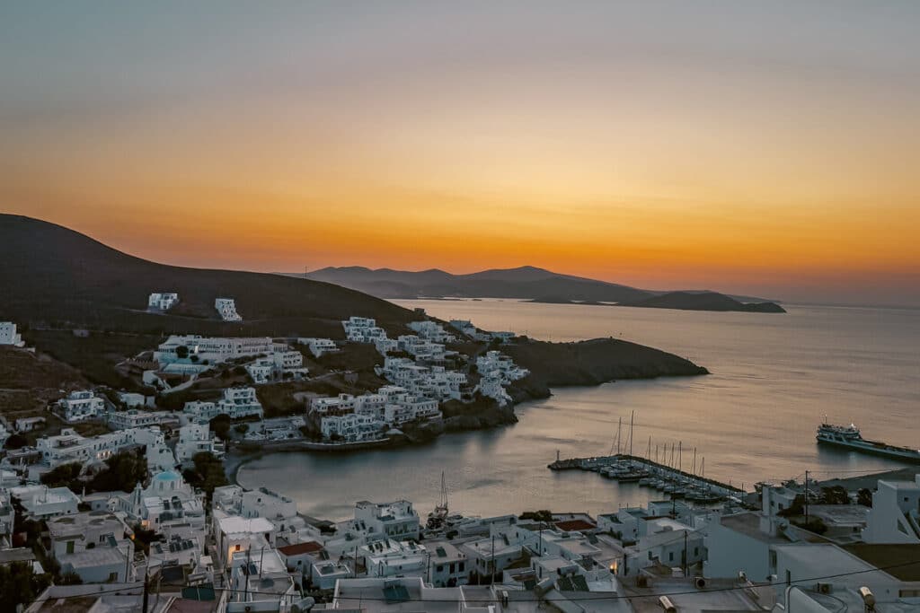 Sunset view over Astypalaia with whitewashed houses, the harbor, boats, surrounding hills, and the Aegean Sea glowing under an orange sky.