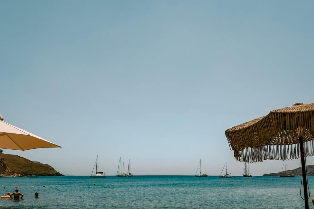 Livadi Beach in Astypalaia with calm turquoise water, swimmers near the shore, sailboats anchored in the bay, and beach umbrellas framing the view.