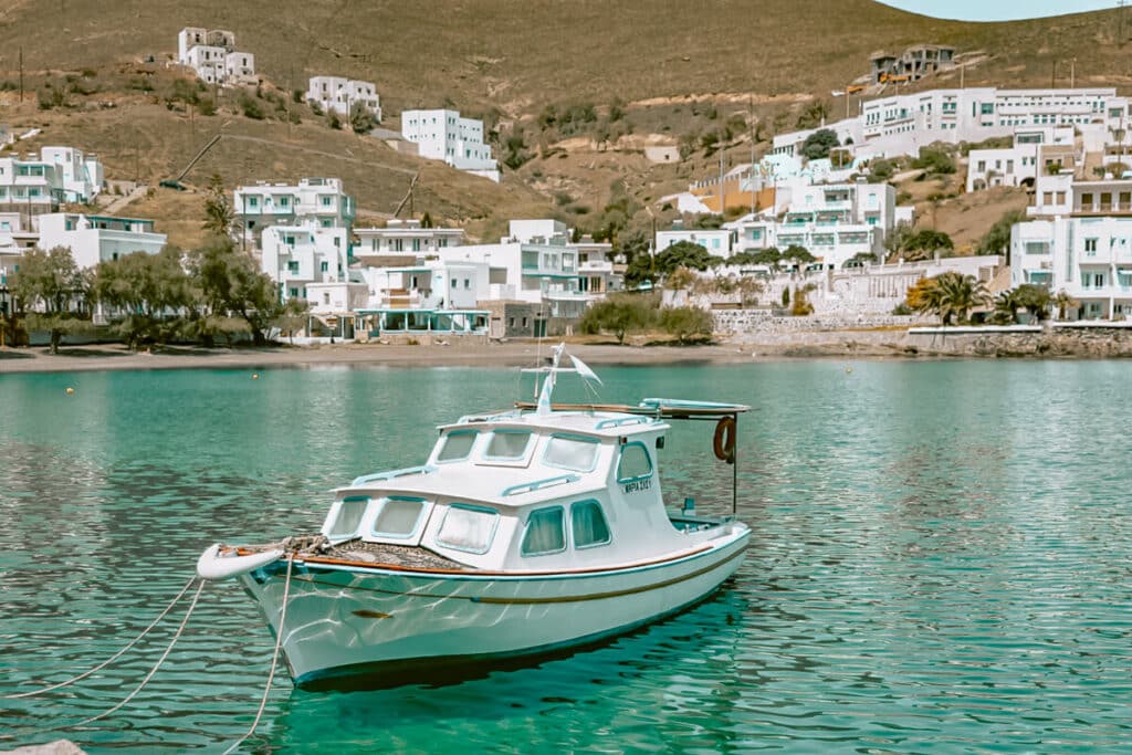 Small white boat floating in turquoise water at Pera Gialos Bay in Astypalaia, with whitewashed buildings and dry hills rising behind the harbor.