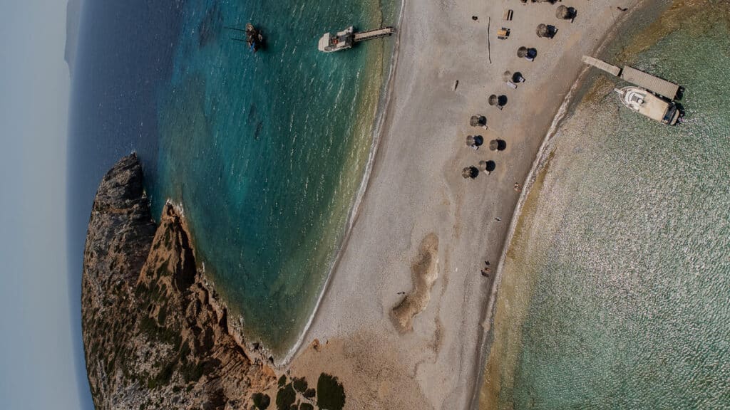 Aerial view of Kounoupa beach in Astypalaia, with a narrow sandy strip between clear turquoise water, small boats, umbrellas, and rocky coastline.